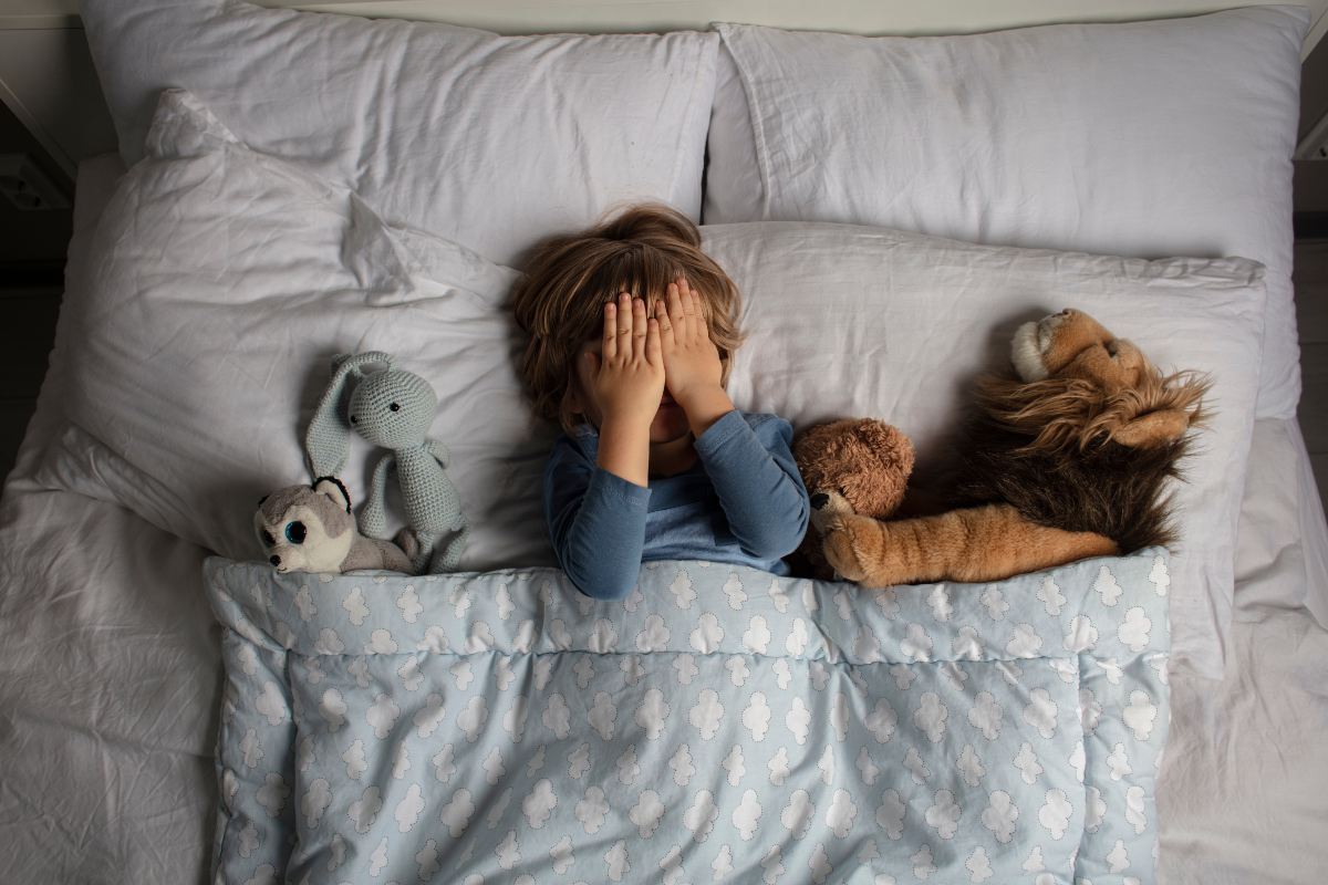 O fotografie cu un băiețel așezat în pat, alături de animale de pluș, folosită pentru a ilustra coșmarurile la copii