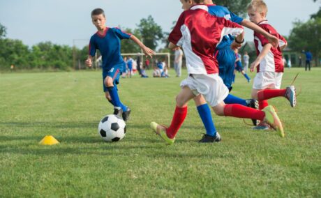 Băieți, îmbrăcați cu echipament sportiv albastru și roșu cu alb, joacă fotbal pe un teren cu iarbă verde ilustrând cum să previi loviturile copiilor, atunci când fac sport în aer liber 