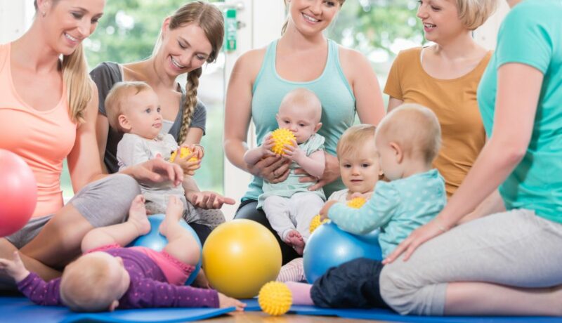 Grup de mame cu bebeluși pe care îi țin în brațe sau stau sprijiniți pe mingi colorate, din cauciuc, mici și mari, și un bebeluș care stă pe spate, pe o saltea albastră, care s-au reunit într-o sală luminoasă, care ilustrează cum găsești un grup de mame