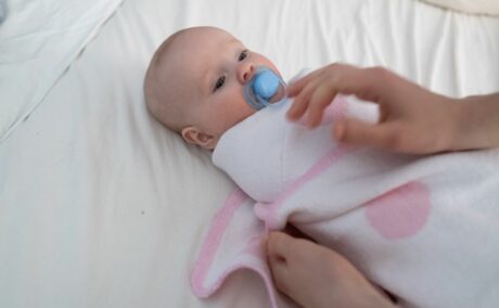 Bebeluș întins pe patul acoperit cu cearșaf alb, care are o suzetă albastră în gură și stă pe spate în timp ce mama îl înfașă cu o păturică alb cu pete mari, roz, ilustrând cum să înfeși bebelușul