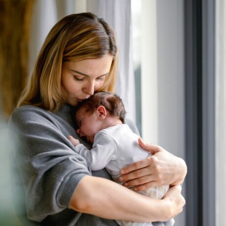 Mămică, îmbrăcată cu o bluză gri, care stă la ferească și ține un nou născut în brațe, ilustrând Cum să te joci cu nou-născutul