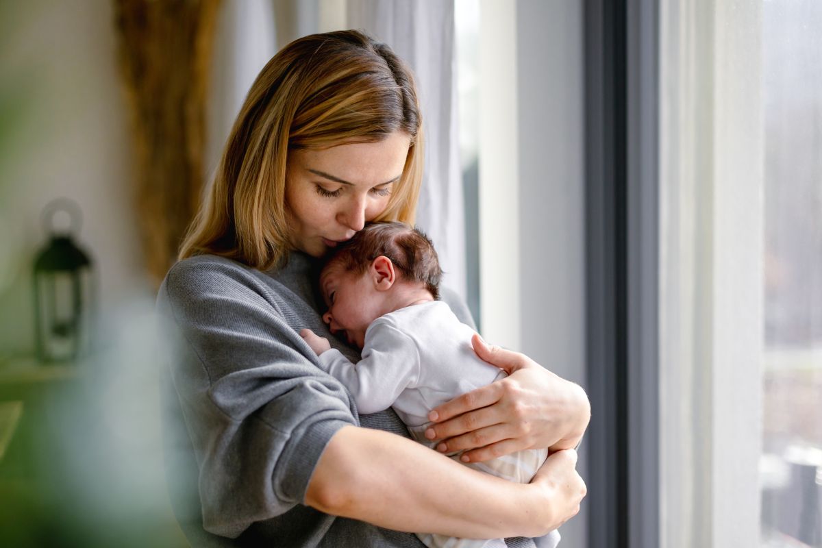 Mămică, îmbrăcată cu o bluză gri, care stă la ferească și ține un nou născut în brațe, ilustrând Cum să te joci cu nou-născutul