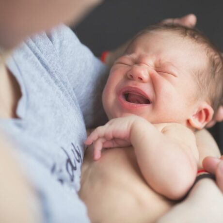 Mămică, îmbrăcată un tricou gri cu text în jurul pieptului, ține în brațe un bebeluș care plânge, ilustrând de ce își arcuiesc bebelușii spatele