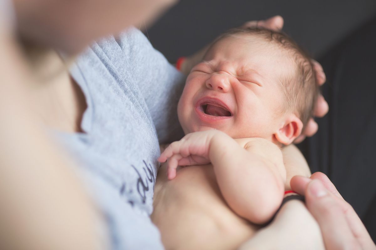 Mămică, îmbrăcată un tricou gri cu text în jurul pieptului, ține în brațe un bebeluș care plânge, ilustrând de ce își arcuiesc bebelușii spatele