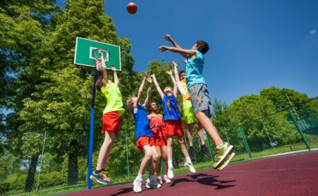 Copii, îmbrăcați colorat, pe un teren de baschet, într-o zonă împădurită, aruncând o minge la coș, ilustrând de ce refuză copiii să facă sport