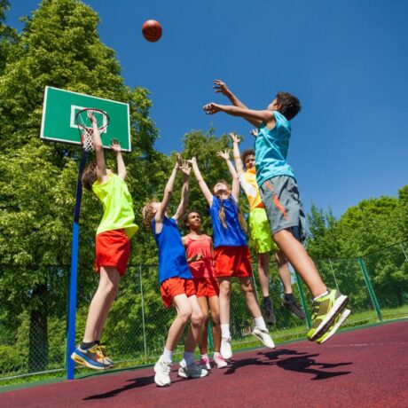 Copii, îmbrăcați colorat, pe un teren de baschet, într-o zonă împădurită, aruncând o minge la coș, ilustrând de ce refuză copiii să facă sport
