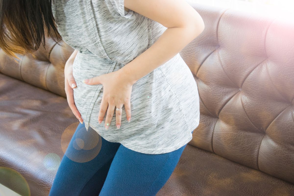 Femeie însărcinată, îmbrăcată cu o bluză gri și jeanși, care stă ușor aplecată în față și își ține burta cu mâinile, sugerând dezlipirea de placentă, iar în stape are o canapea din piele, maro