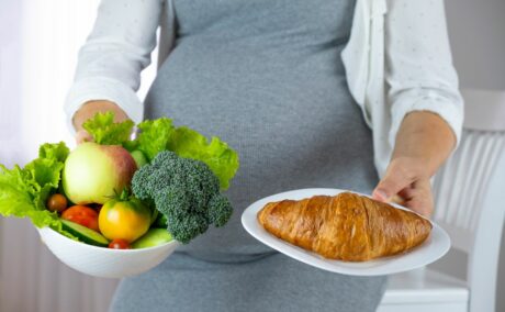 Femeie însărcinată, îmbracată cu o rochie gri, mulată, și o cămașă abă cu puncte bleu, pe deasupra, care ține în mână un castron cu legume și în cealaltă o farfurie cu un croasant, ilustrând diabetul gestațional