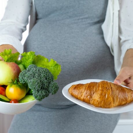Femeie însărcinată, îmbracată cu o rochie gri, mulată, și o cămașă abă cu puncte bleu, pe deasupra, care ține în mână un castron cu legume și în cealaltă o farfurie cu un croasant, ilustrând diabetul gestațional