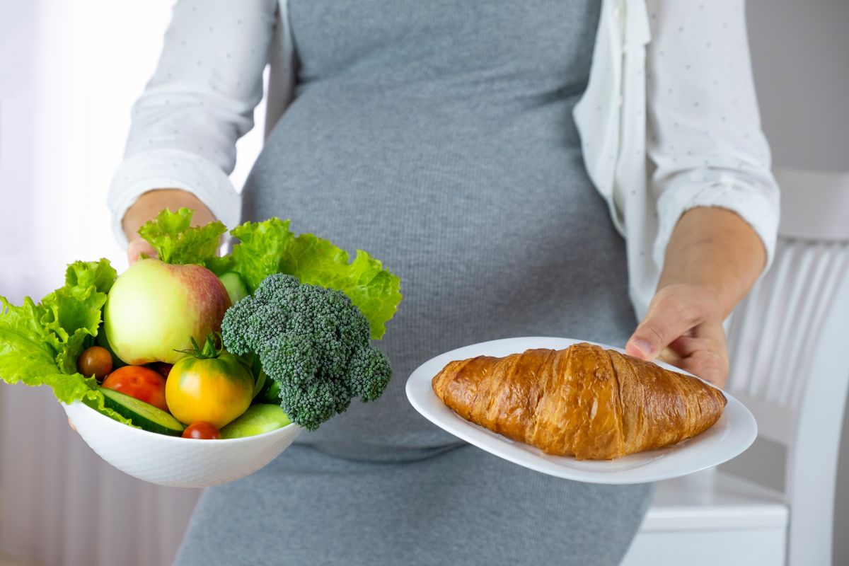 Femeie însărcinată, îmbracată cu o rochie gri, mulată, și o cămașă abă cu puncte bleu, pe deasupra, care ține în mână un castron cu legume și în cealaltă o farfurie cu un croasant, ilustrând diabetul gestațional