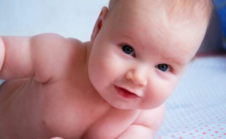 Bebeluș dezbrăcat, așeazat pe pat, pe un cearșaf alb cu buline roșii, pus pe burtă, sprijinit pe un braț, ilustrând distocia de umăr