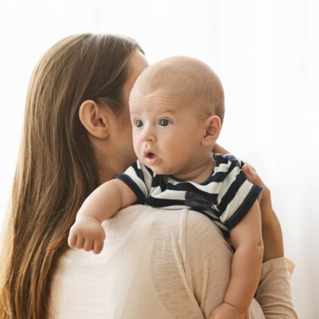 Mamă care stă cu spatele și ține pe umăr un bebeluș, care are un tricou alb cu bleumarin, ilustrând eructația la bebeluși