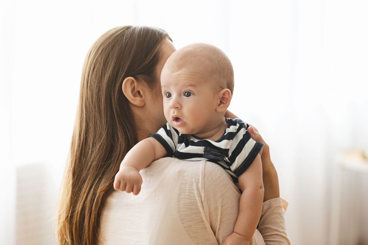 Mamă care stă cu spatele și ține pe umăr un bebeluș, care are un tricou alb cu bleumarin, ilustrând eructația la bebeluși