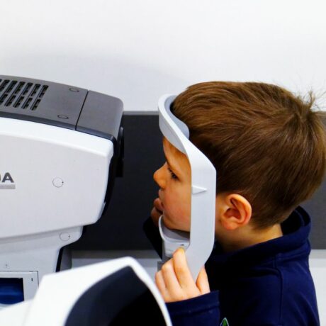Băiețel, îmbrăcat cu o bluză bleumarin, care stă cu capul într-un suport al unui aparat care face teste oftalmologice, ilustrând examenul oftalmologic la copii