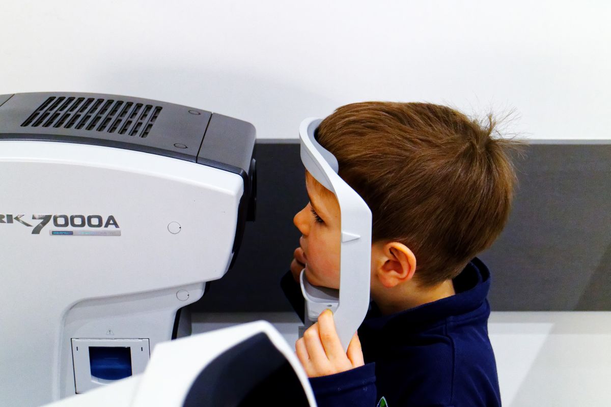 Băiețel, îmbrăcat cu o bluză bleumarin, care stă cu capul într-un suport al unui aparat care face teste oftalmologice, ilustrând examenul oftalmologic la copii