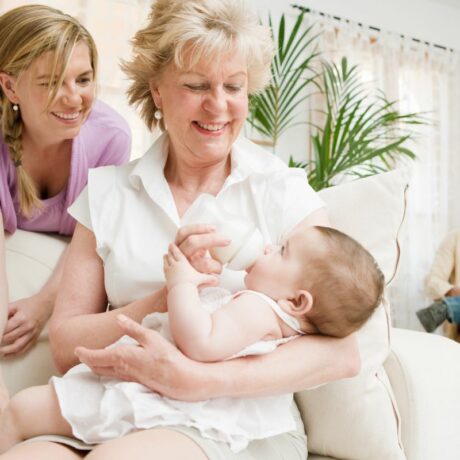 Bunică, îmbrăcată cu o bluză albă cu mâneci scurte, în vizită la un nou-născut, pe care îl ține în brațe, în timp ce îi oferă un biberon, stând pe o canapea bej, iar mama bebelușului stă sprijinită pe canapea și privește, iar tatăl stă pe un scaun