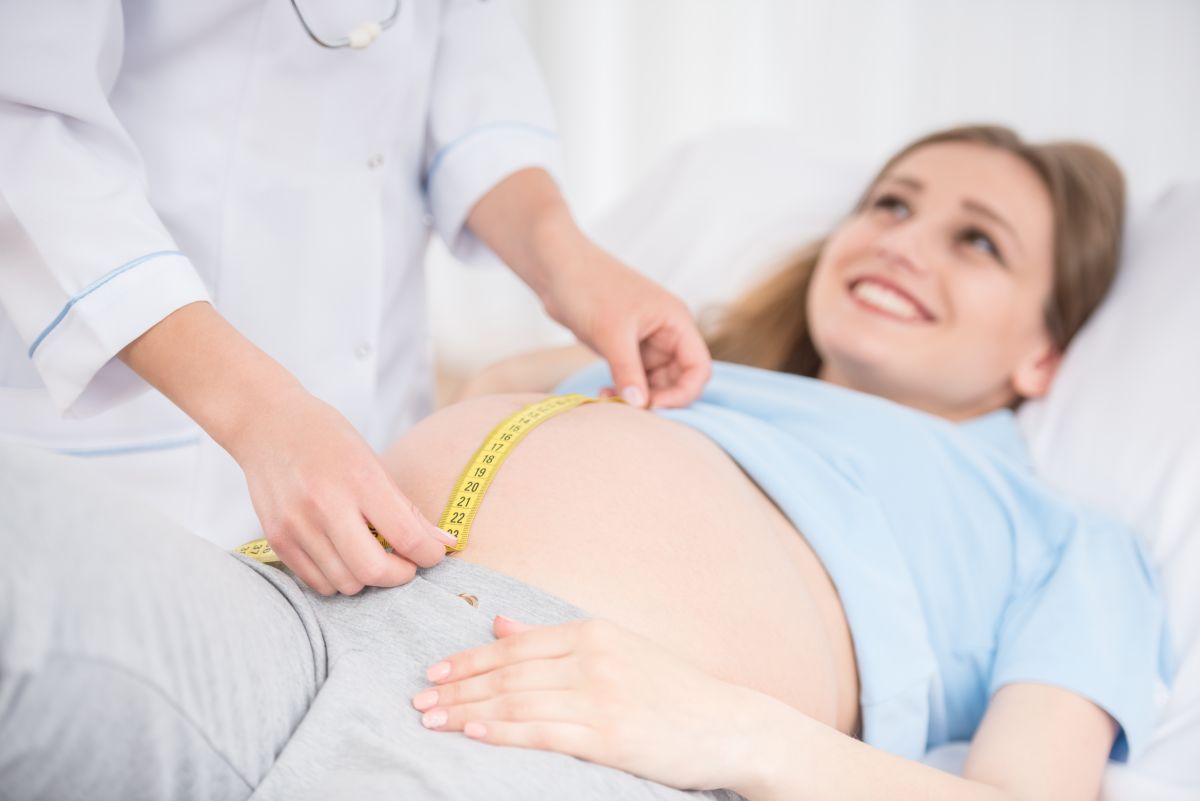 Femeie însărcinată, care stă întinsă pe spate, îmbrăcată cu colanți gri și bluză cu mâneci scurte, cu burta descoperită și medic în halat alb, care măsoară înălțimea fundului uterin cu un centimetru galben