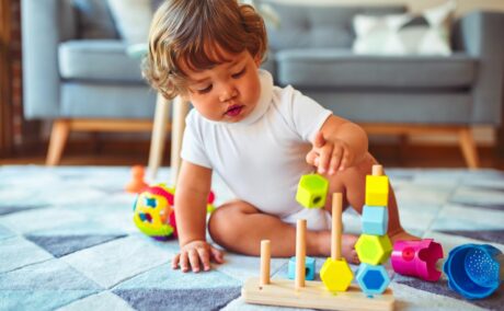 Toddler care se joacă pe un covor cu romburi bleu, care se joacă cu niște înșirând hexagoane din lemn, pe un suport, iar în spate are o canapea bleu, ilustrând încurajează toddlerul să se joace independent