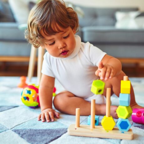 Toddler care se joacă pe un covor cu romburi bleu, care se joacă cu niște înșirând hexagoane din lemn, pe un suport, iar în spate are o canapea bleu, ilustrând încurajează toddlerul să se joace independent