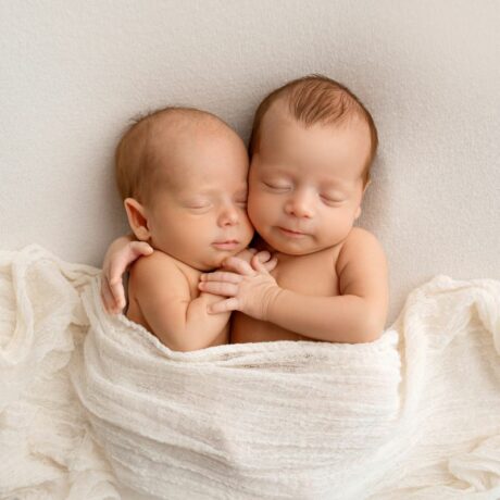 Bebeluși gemeni, care stau îmbrățișați, așezați pe pat și acoperiți cu o păturică albă, până la mijloc, ilustrând lucruri surprinzătoare despre gemeni