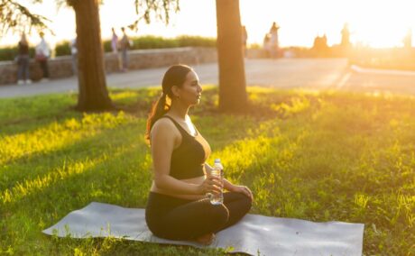 Femeie însărcinată, îmbrăcată cu colanți și bustieră maro, care face yoga, ca una dintre metode de a crește fertilitatea natural, pe o saltea albă, pe iarbă, într-un parc și ține în mână o sticlă cu apă,