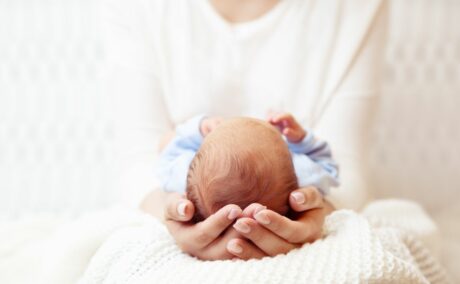 Mamă care își ține bebelușul, îmbrăcat cu un body alb cu bleu, pe genunchi, pe o pătură tricotată, albă, cu picioarele spre abdomenul ei, cuprinzându-i capul cu mâinile, ilustrând microcefalia.