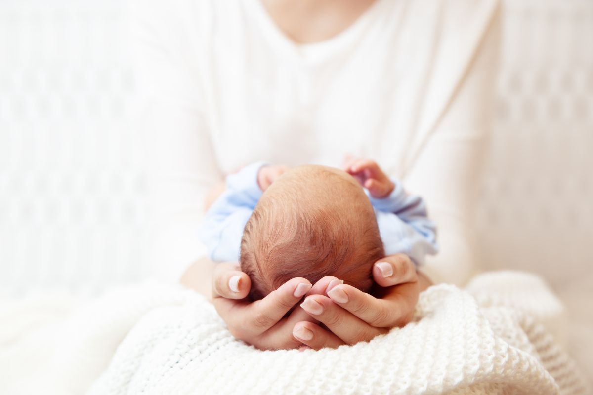 Mamă care își ține bebelușul, îmbrăcat cu un body alb cu bleu, pe genunchi, pe o pătură tricotată, albă, cu picioarele spre abdomenul ei, cuprinzându-i capul cu mâinile, ilustrând microcefalia.