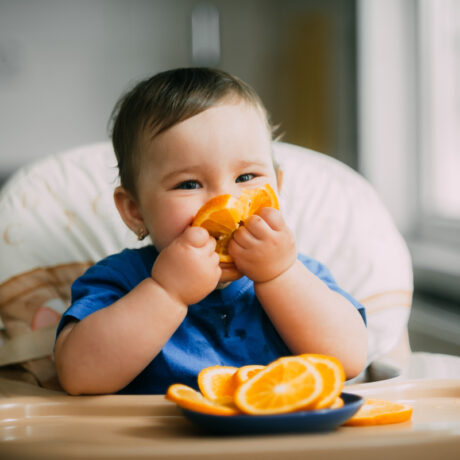 O fotografie cu un bebeluș care mănâncă felii de portocală, așezat într-un scaun de masă, folosită pentru a ilustra nevoile nutriționale ale bebelușilor