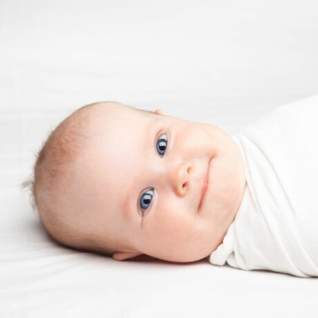 Bebeluș cu ochi albaștri, înfășat într-o pânză albă, care stă cu capul într-o parte, așezat pe patul acoperit cu cearșaf alb, care ilustrează până când poți înfășa bebelușul