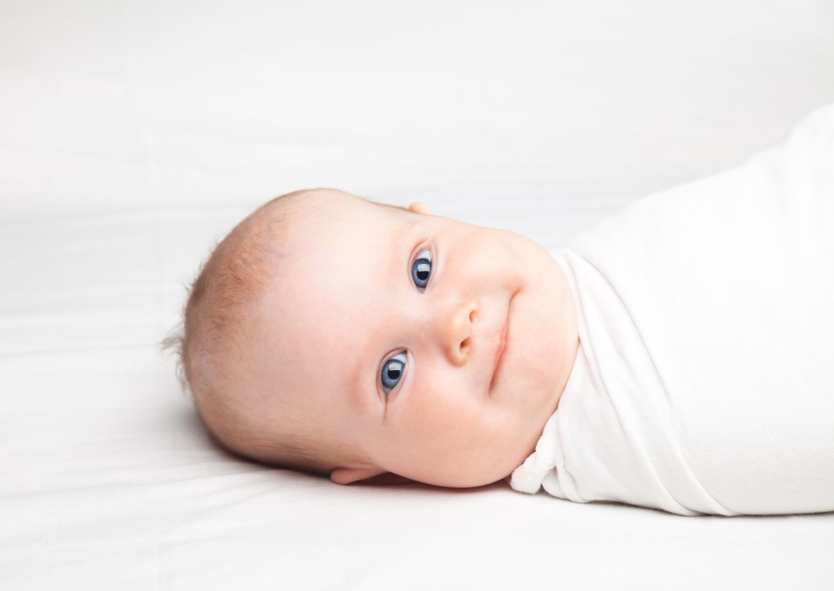 Bebeluș cu ochi albaștri, înfășat într-o pânză albă, care stă cu capul într-o parte, așezat pe patul acoperit cu cearșaf alb, care ilustrează până când poți înfășa bebelușul