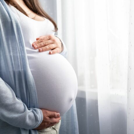 Femeie însărcinată în ultimul trimestru de sarcină, care are polihidramnios, îmbrăcată cu pantaloni bej, maiou alb și cardigan bleu, care stă în dormitor, lângă ferestra cu perdea albă