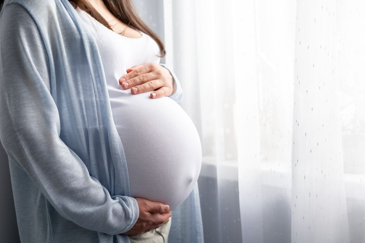 Femeie însărcinată în ultimul trimestru de sarcină, care are polihidramnios, îmbrăcată cu pantaloni bej, maiou alb și cardigan bleu, care stă în dormitor, lângă ferestra cu perdea albă