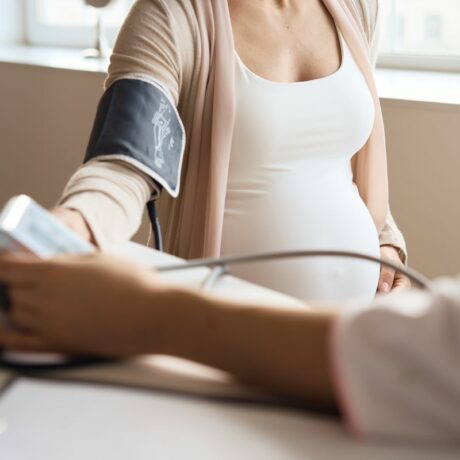 Femeie însărcinată, îmbrăcată cu maiou alb și cardigan bej, care se confruntă cu preeclampsia și a venit în cabinetul medicului pentru a-i verifica tensiunea cu aparat de luat tensiunea pe care i l-a pus pe braț