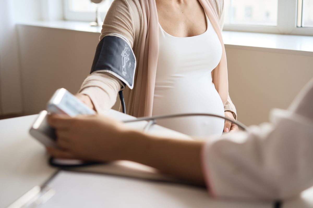 Femeie însărcinată, îmbrăcată cu maiou alb și cardigan bej, care se confruntă cu preeclampsia și a venit în cabinetul medicului pentru a-i verifica tensiunea cu aparat de luat tensiunea pe care i l-a pus pe braț