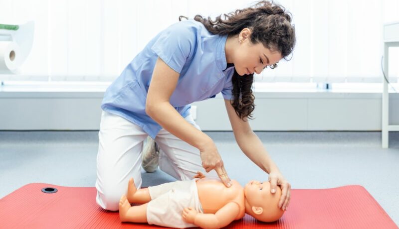 Medic îmbrăcat cu bluză albastră șoi pantaloni albi, care stă în genunchi, pe o saltea roșie și explică pe o păpușă cu se acordă primul ajutor pediatric