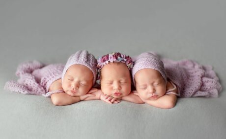 Tribleți care dorm, așezați pe abdomen cu mâinile sub bărbie, acoperiți cu o pătru tricotată mov deschis și pe cap doi dintre ei au căciuli și bebelușul din mijloc are pe cap o bentiță cu flori mov și roz, ilustrând sarcina cu tripleți