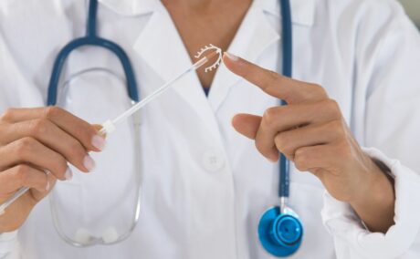Medic în halat alb, care are de gât un stetoscop albastru și ține într-o mână steriletul și degetul arătător de la mâna cealaltă atinge vârful
