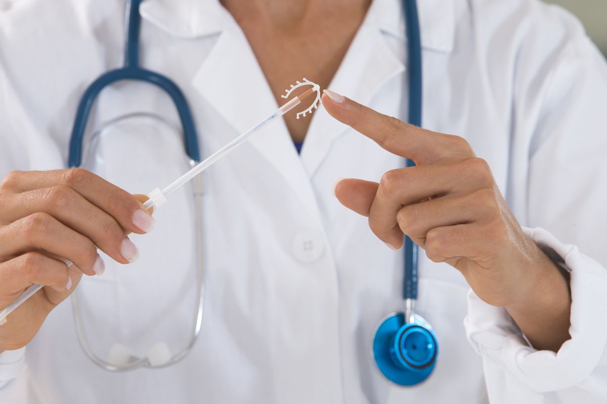 Medic în halat alb, care are de gât un stetoscop albastru și ține într-o mână steriletul și degetul arătător de la mâna cealaltă atinge vârful