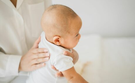 Medic îmbrăcat în halat alb, care ține un bebeluș. îmbrăcat cu body alb, cu mâneci scurte, pe genunchi, și îl mângâie ușor pe spate pentru a îndepărta sughițul bebelușului