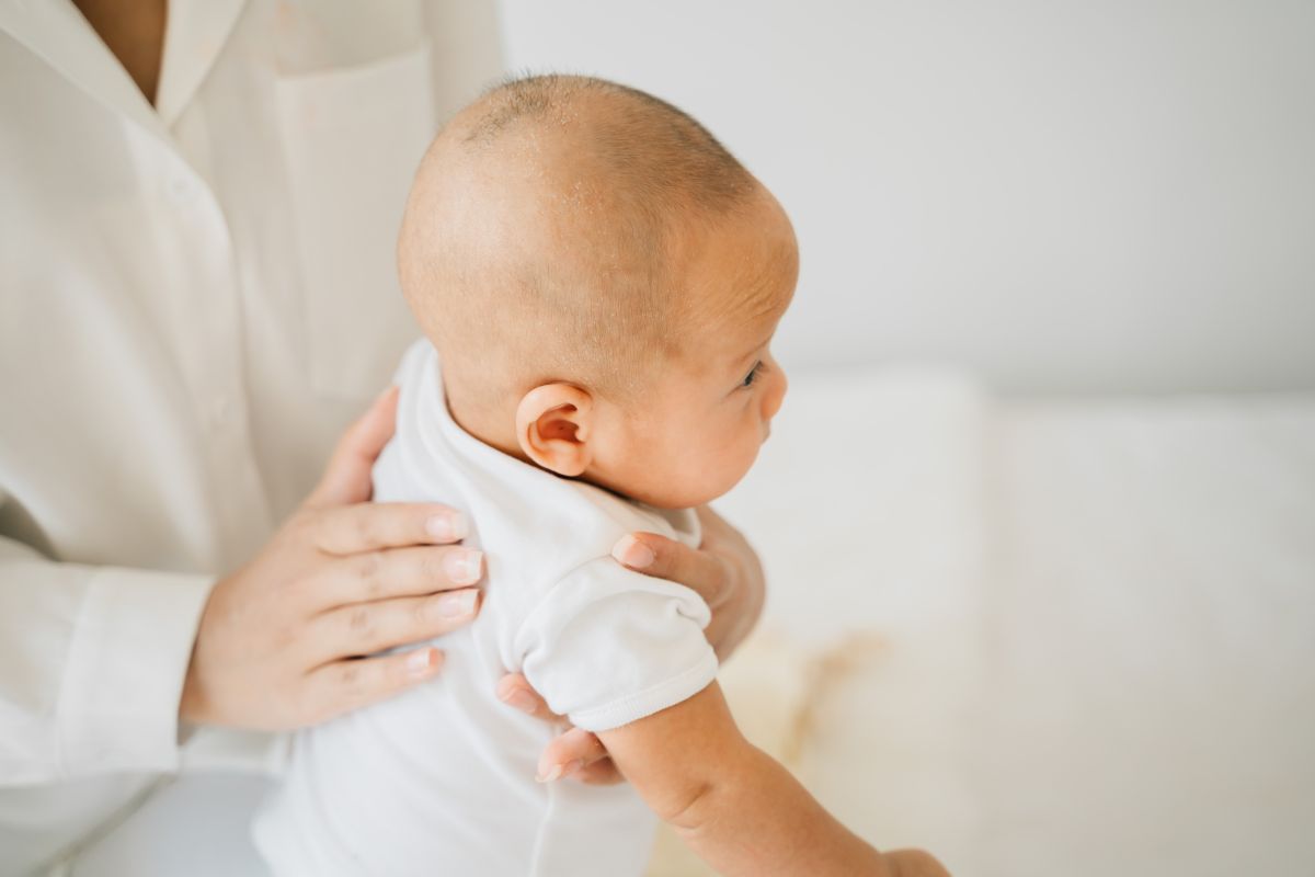 Medic îmbrăcat în halat alb, care ține un bebeluș. îmbrăcat cu body alb, cu mâneci scurte, pe genunchi, și îl mângâie ușor pe spate pentru a îndepărta sughițul bebelușului