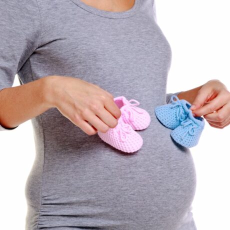 Femeie însărcinată, îmbrăcată într-o bluză gri, care ține într-o mână botoșei roz și în cealaltă botoșei bleu, ilustrând superstiții legate de prezicerea sexului copilului