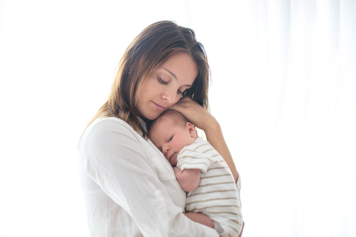 Mamă, îmbrăcată cu cămașă albă, cu un nou-născut în brațe, îmbrăcat într-un body alb cu dungi bej, ilustrând trimestrul 4 de sarcină