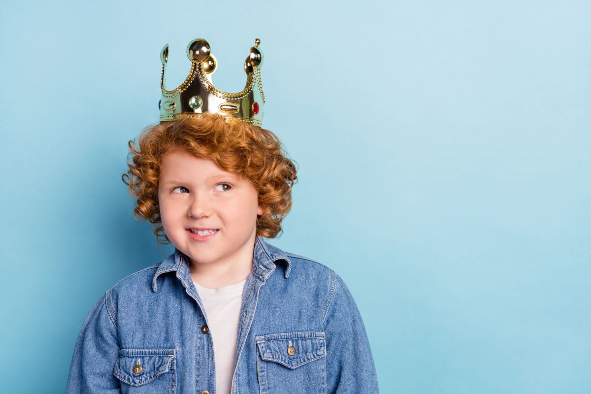 Băiețel roșcat, cu tricou alb și jachetă din denim, pe un fundal bleu, care are pe cap o coroană de rege, ilustrând 3 greșeli de parenting care îi fac pe copii narcisiști