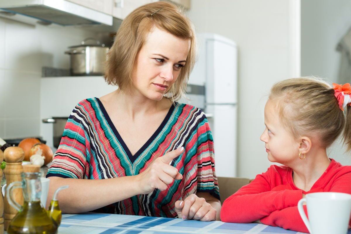 Mamă, îmbrăcată cu o bluză cu dungi colorate, care stă pe scaun, în bucătărie, în fața unei mese acoperite cu dungi și pătrate albastre și arată cu degetul către fiica ei, îmbrăcată cu o bluză roșie, ilustrând 5 tipuri de reguli de care copiii au nevoie