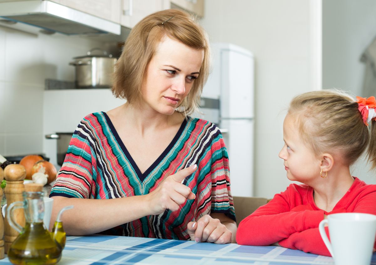 Mamă, îmbrăcată cu o bluză cu dungi colorate, care stă pe scaun, în bucătărie, în fața unei mese acoperite cu dungi și pătrate albastre și arată cu degetul către fiica ei, îmbrăcată cu o bluză roșie, ilustrând 5 tipuri de reguli de care copiii au nevoie