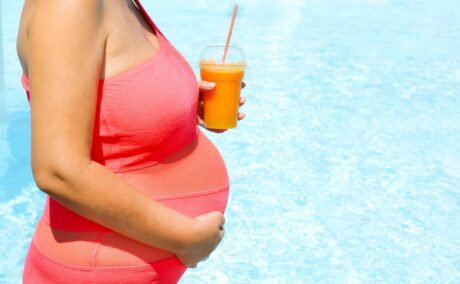 Femeie însărcinată, lângă piscină, îmbrăcată cu un costum de baie roz, care ține în mână un pahar din plastic cu suc de portocale, ilustrând supraîncălzirea în sarcină