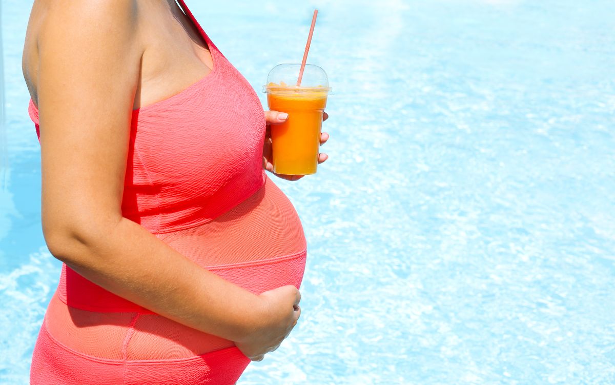 Femeie însărcinată, lângă piscină, îmbrăcată cu un costum de baie roz, care ține în mână un pahar din plastic cu suc de portocale, ilustrând supraîncălzirea în sarcină