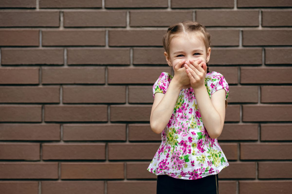 Fetiță, îmbrăcată cu o bluză cu flori roz și verz, stă lângă un zid și își ține mâinile la gură, ilustrând copilul înjură