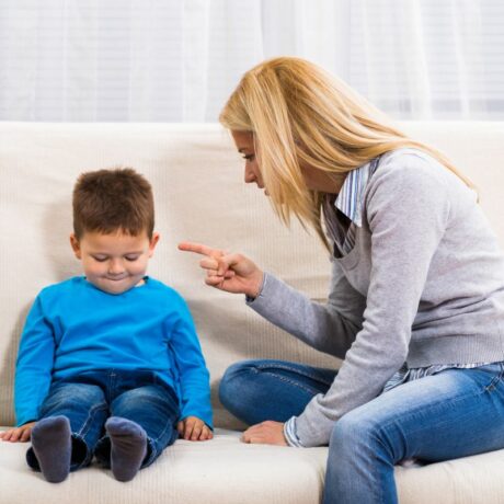 Mamă, îmbrăcată cu o cămașă cu dungi albastre și verzi, cu un pulover gri pe deasupra și jeanși, care stă pe o canapea bej, îndreptată și arătând cu degetul arătător către băiețelul ei, îmbrăcat cu pulover albastru și jeanși, ilustrând consecințele folosite în educația copilului