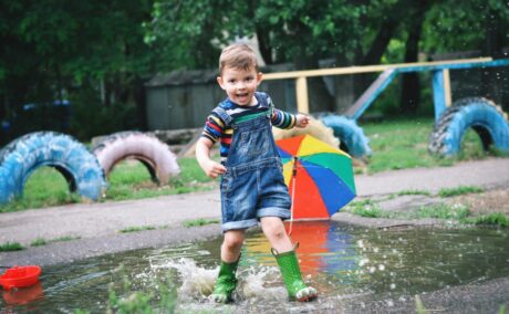 Băiețel, îmbrăcat cu o bluză cu dungi colorate și o salopetă din denim albastru, cu pantaloni scurți și cizme verzi, din cauciuc, care aleargă într-o baltă, în parc, ilustrând consecințele naturale în educația copiilor